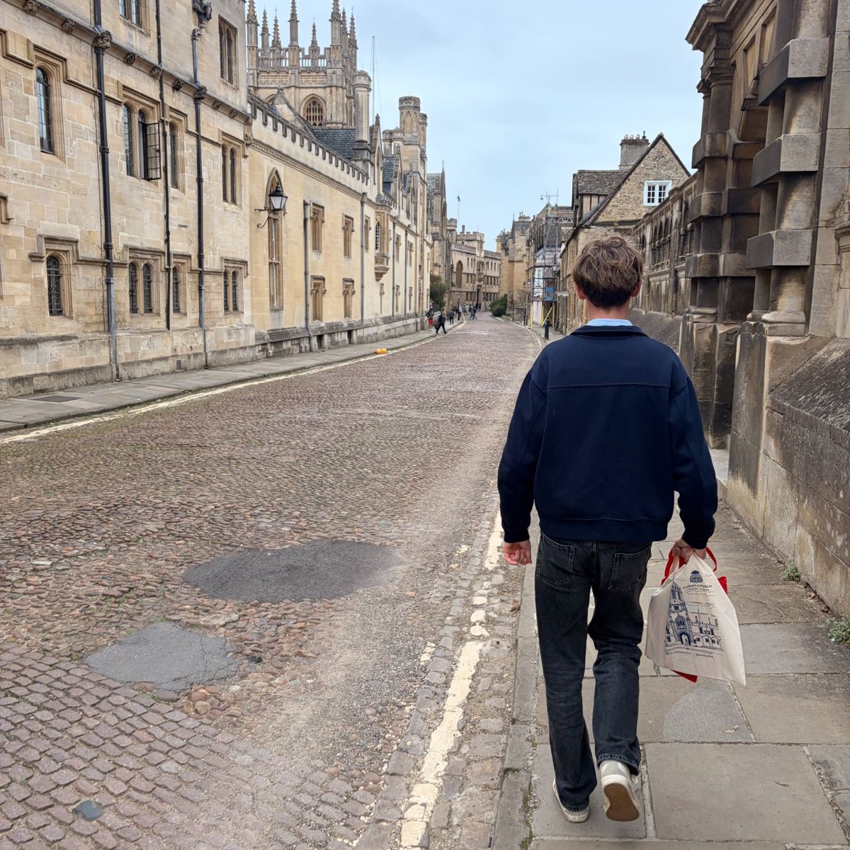 A teenager walking away from the camera up a cobbled Oxford street on a university open day, carrying a college tote bag.